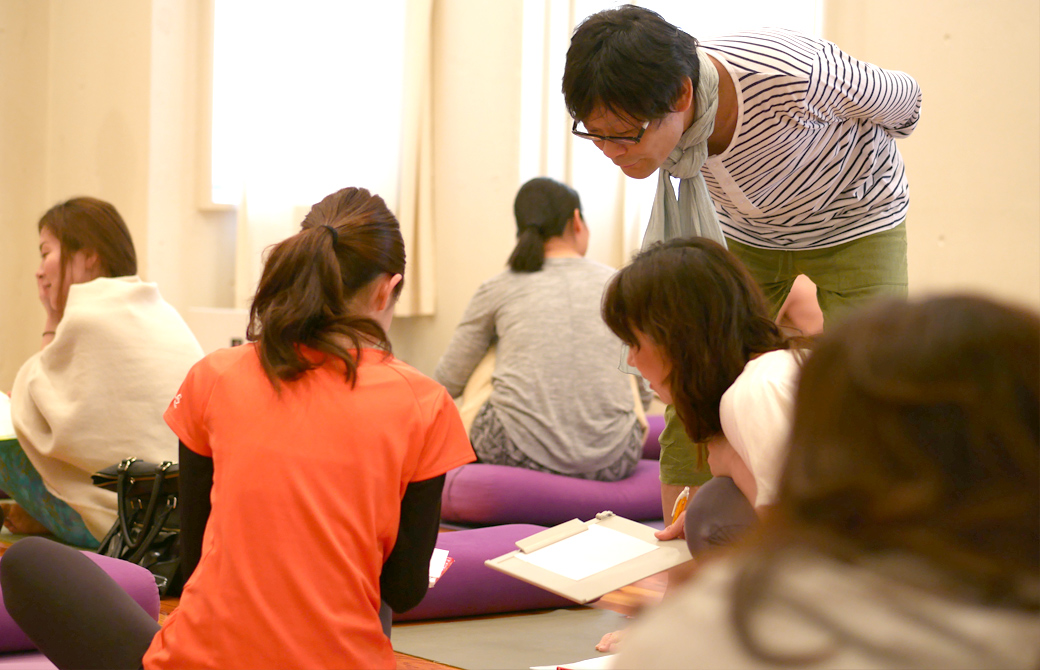 ヨガ×東洋医学の講座風景。講師内田かつのりがグループワーク中の生徒をのぞき込んで見守っている。