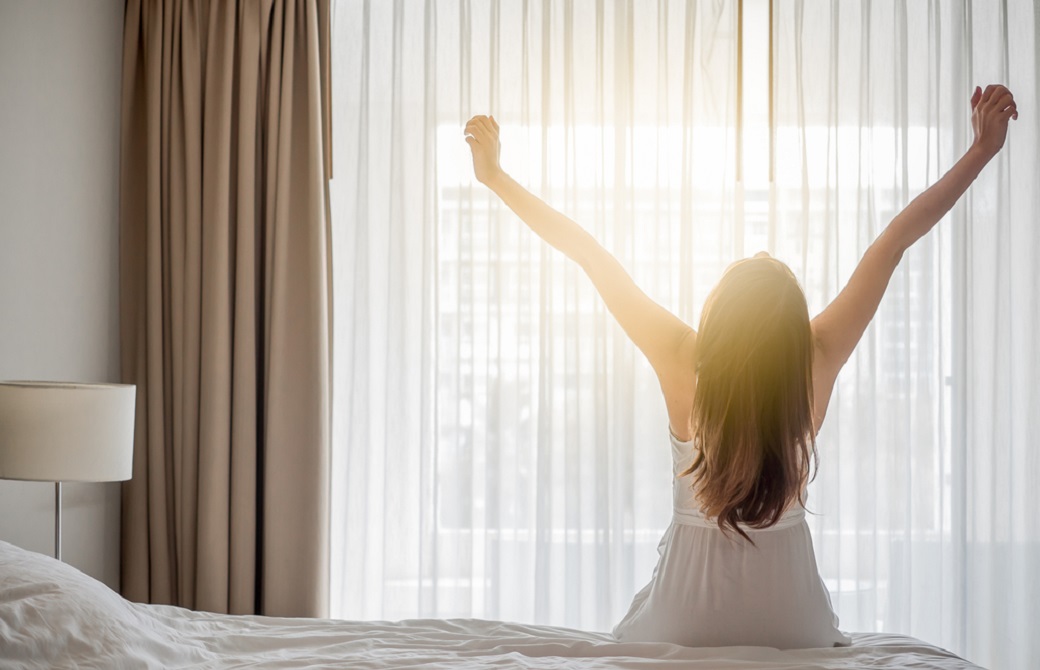 女性がベッドに座り朝日を浴びながら伸びをしている後ろ姿