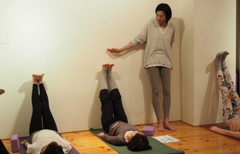 仰向けで壁に足を上げる生徒さんの足に手をかざすサントーシマ香先生