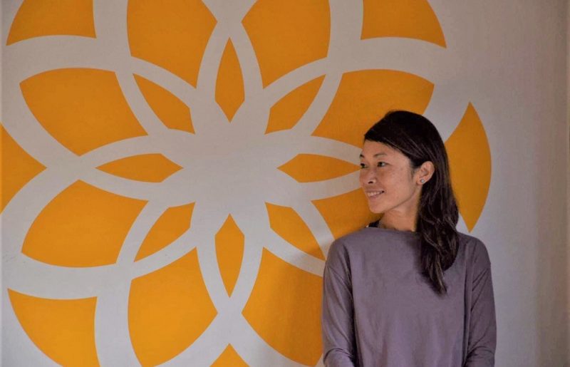 semperviva yoga studioの黄色い花模様の壁の前で横を向いて微笑むマイコ先生