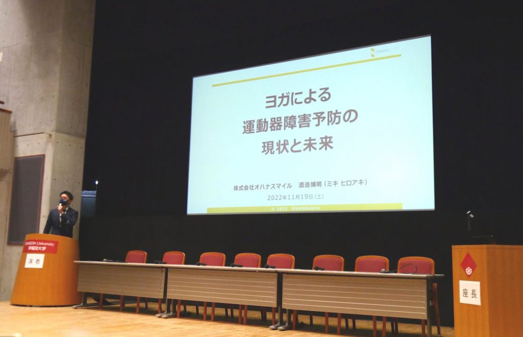 大きなモニターの横に立ち、マイクを持ち話しているMIKIZO